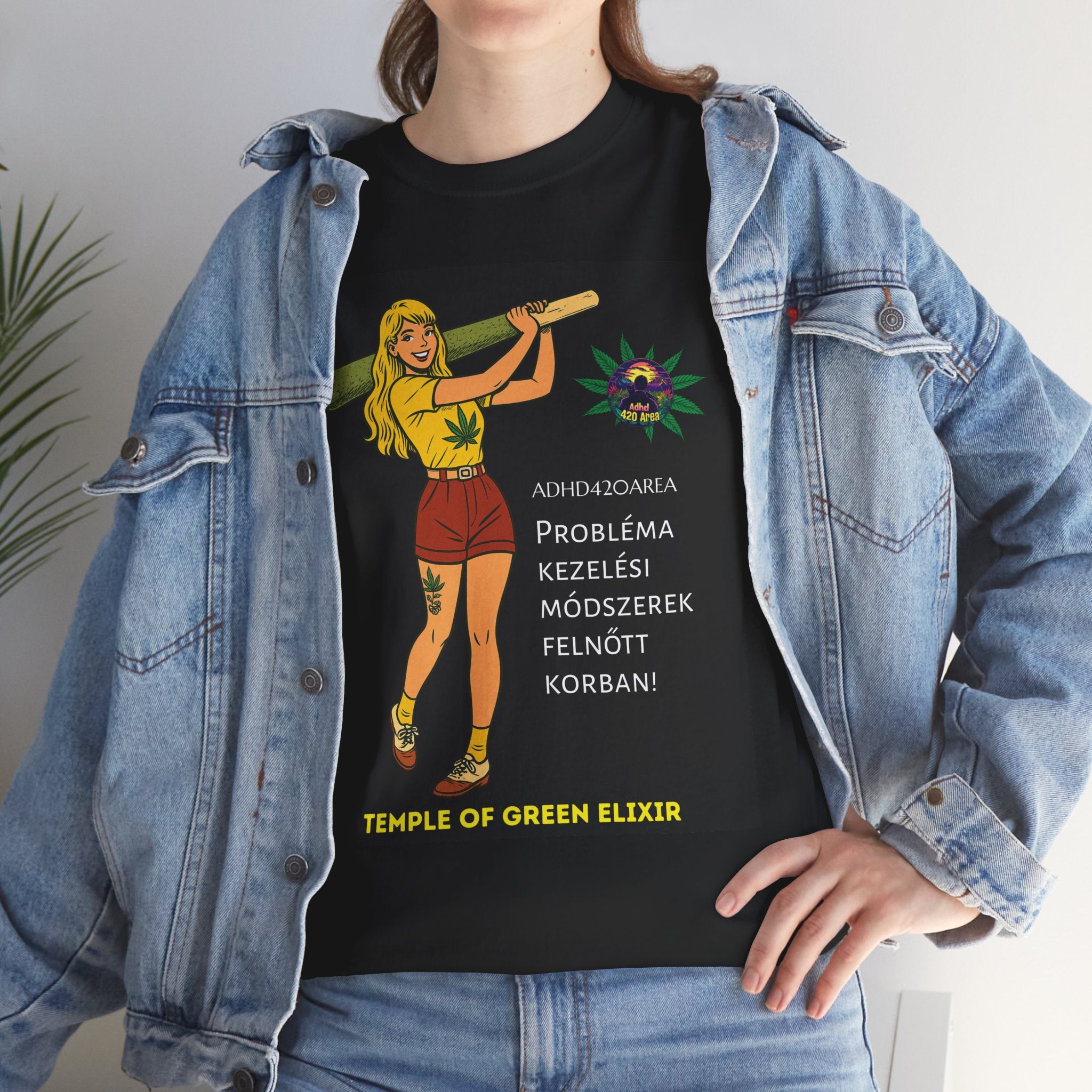 Junge Frau trägt ein schwarzes T-Shirt mit einer Grafik einer gezeichneten Figur, die eine große grüne Zigarette hält. Daneben sind Hanfblätter und der Text auf Deutsch: „ADHD42QAREA Problembehandlungsmethoden im Erwachsenenalter! Temple of Green Elixir“. Sie steht vor weißem Hintergrund und trägt eine blaue Jeansjacke.