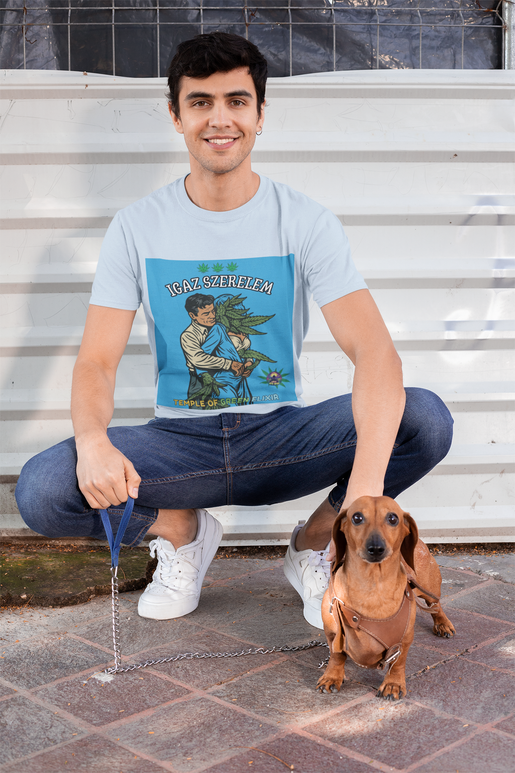 Ein junger Mann posiert lächelnd in der Hocke neben seinem Dackel. Er trägt das hellblaue Wahre Liebe T-Shirt, dessen satirisches Retro-Motiv – die Umarmung eines Mannes mit einer Cannabis-Frau – einen humorvollen Akzent setzt. Sein entspannter Alltagslook mit Jeans und weißen Sneakern zeigt, wie vielseitig das Shirt kombinierbar ist.﻿