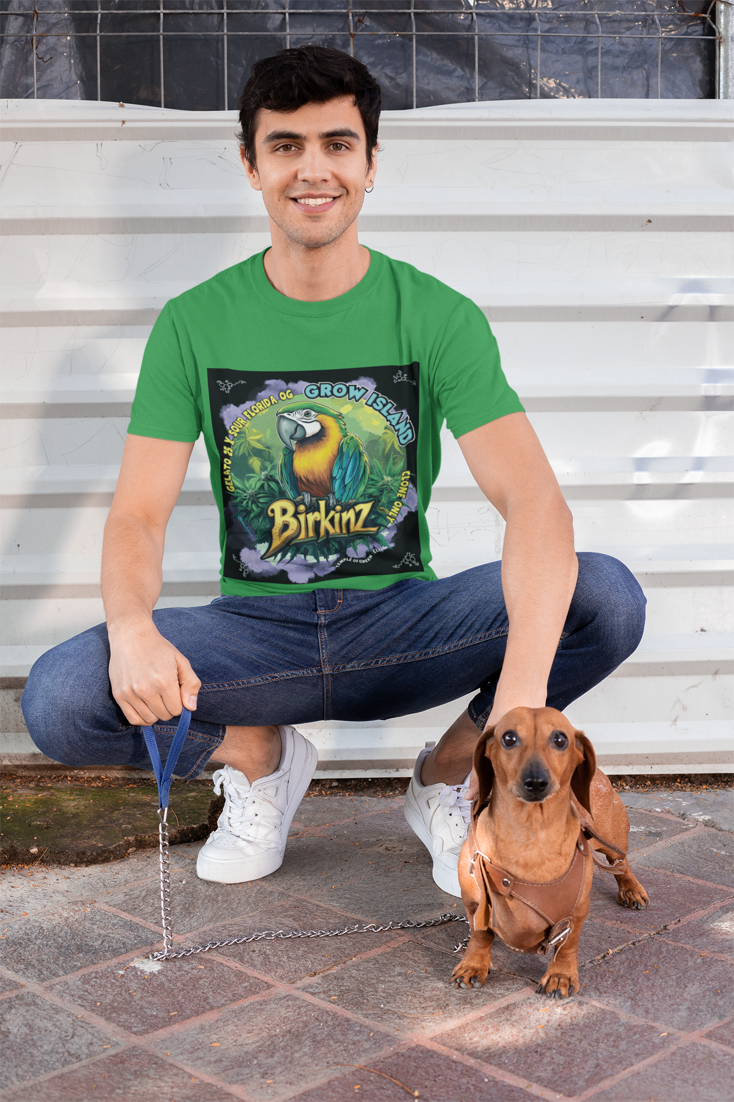 Model hockt neben seinem braunen Hund in einem Birkinz irish grün T-shirt mit blau-grün-gelb Papagei und cannabis blühten rund herum
