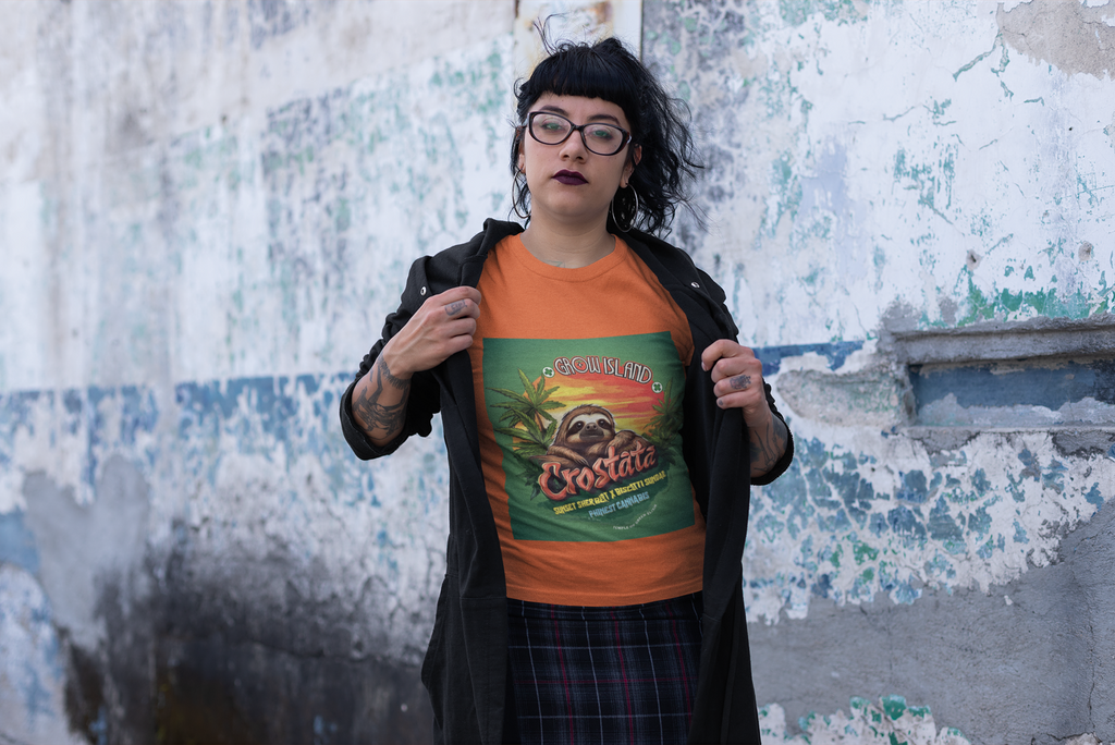 Eine schwarzhaarige, tatowierte, Brillenträgerin Hipsterin steht auf der Straße, trägt einen karierten Rock und einen schwarzen offenen Pullover, unter dem ein oranges Crostata T-Shirt sichtbar ist. Auf dem T-Shirt ist ein Faultier, das im goldenen Licht des Sonnenuntergangs ruht und voller Zufriedenheit auf das duftende Cannabisfeld um sich herum blickt. 