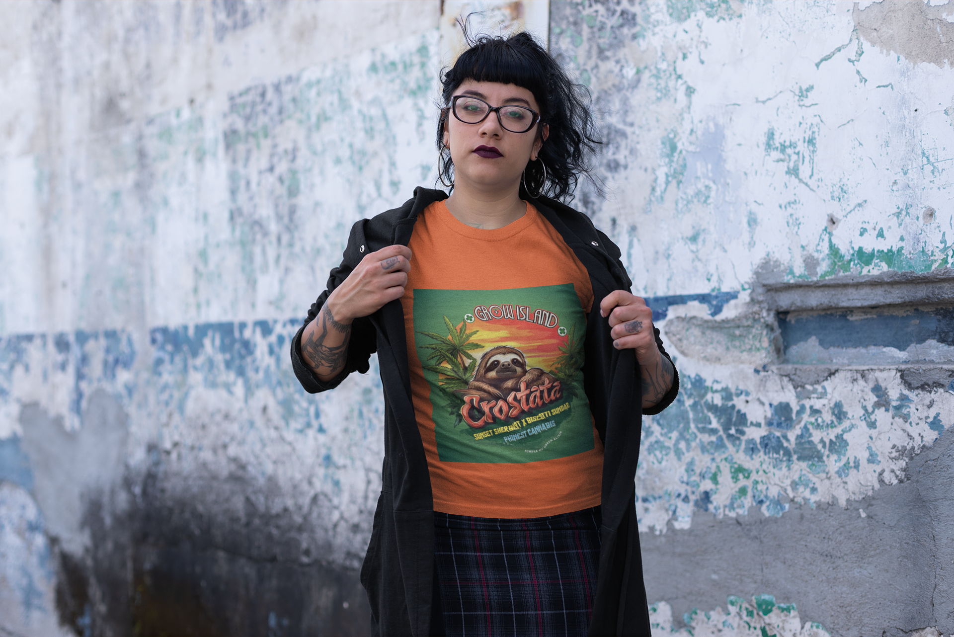 Eine schwarzhaarige, tatowierte, Brillenträgerin Hipsterin steht auf der Straße, trägt einen karierten Rock und einen schwarzen offenen Pullover, unter dem ein oranges Crostata T-Shirt sichtbar ist. Auf dem T-Shirt ist ein Faultier, das im goldenen Licht des Sonnenuntergangs ruht und voller Zufriedenheit auf das duftende Cannabisfeld um sich herum blickt. 