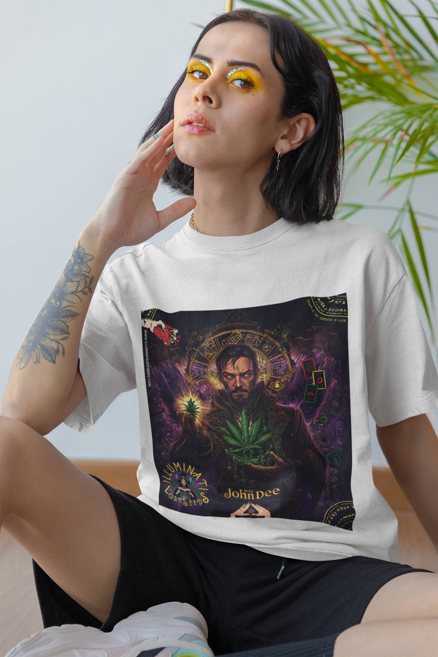 Eine junge Frau mit auffälligem gelbem Augen-Make-up und einem Tattoo am Arm präsentiert das weiße John Dee T-Shirt von Temple of Green Elixir. Ihr intensiver, direkter Blick fesselt den Betrachter und bildet einen spannenden Kontrast zum mystischen und detailreichen Design des Shirts. Diese Kollaboration von "Temple of Green Elixir" und "Illuminati Lost Seeds" ist für selbstbewusste Persönlichkeiten gemacht, die Kunst und Mode als Ausdruck ihrer selbst sehen.﻿