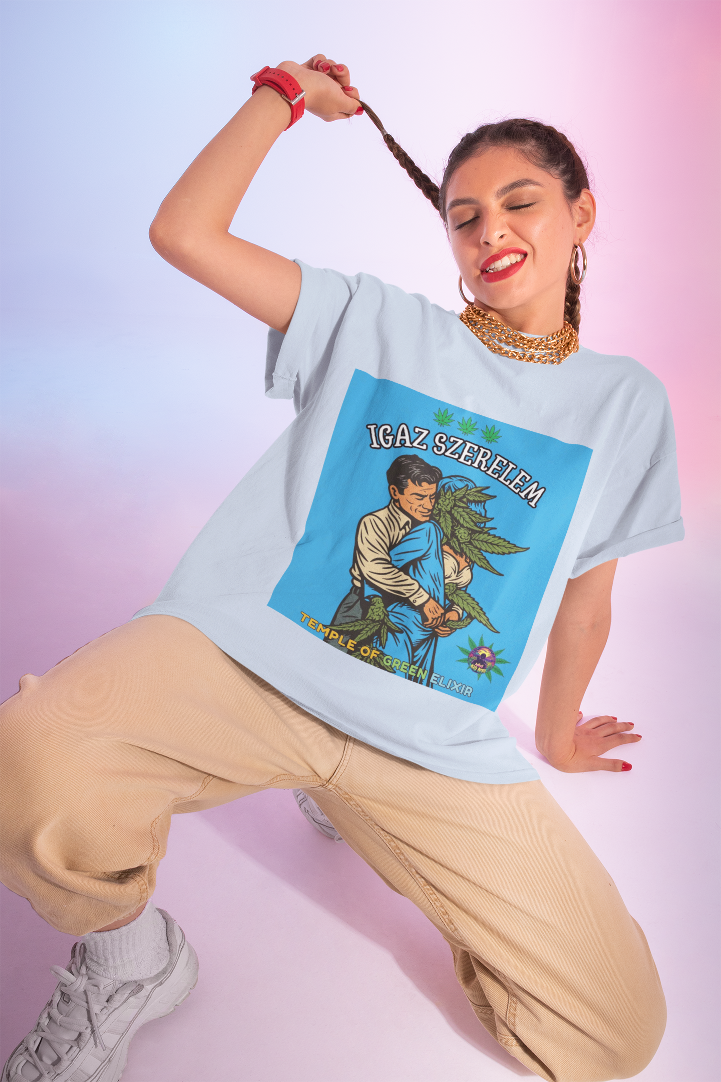 Eine junge Frau mit verspielten Zöpfen und goldenen Ketten kombiniert das hellblaue Wahre Liebe T-Shirt mit dem humorvollen 'Igaz Szerelem'-Motiv zu einer lässigen beigen Hose. Ihr selbstbewusster, fröhlicher Ausdruck unterstreicht perfekt die ironische Botschaft des Designs, das eine liebevolle Umarmung zwischen einem Mann und einer Cannabis-Frau im Retro-Stil zeigt.﻿