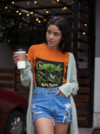 Dame steht mit Kaffebechem im Hand auf der Strasse in orangen Gary Payton & Jealousy T-shirt. Auf dem T-shirt  Zwei mächtige, grün beschuppte Schlangen winden sich umeinander, in ihren Augen funkeln uralte Weisheit und spielerische Herausforderung.  Im Hintergrund rahmen üppige Cannabisblätter die Szene ein und verleihen der Komposition Stärke, Tiefe und eine dunkle Eleganz. 