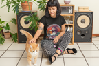 Ein schwarzhaariges Mädchen sitzt auf dem Boden vor Lautsprechern, streichelt ihre rote Katze und trägt ein schwarzes High Society T-Shirt, auf dem Folgendes zu sehen ist: Am Horizont, in den Rot- und Orangetönen des Sonnenuntergangs, nähert sich ein altes Segelschiff, während am Ufer eine Meeres-Sirene mit langem, feuerrotem Haar und spitzen Ohren steht – in ihrem Blick liegen alle Geheimnisse des Meeres. Neben ihrem Arm symbolisieren Cannabisblätter und feiner Rauch die Genüsse des Lebens und das Gefühl.