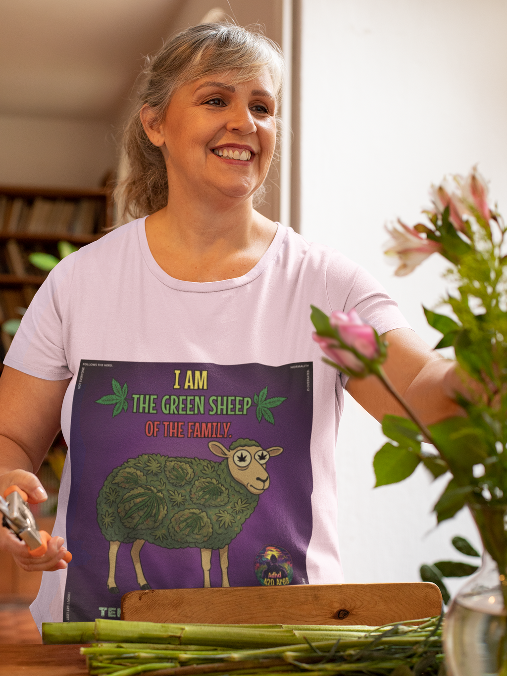 Eine lächelnde, ältere Frau mit grau-blonden Haaren arrangiert einen Blumenstrauß. Sie trägt ein hellviolettes T-Shirt von Temple of Green Elixir mit dem 'I am the Green Sheep of the Family'-Aufdruck. Die Szene ist hell und freundlich.﻿