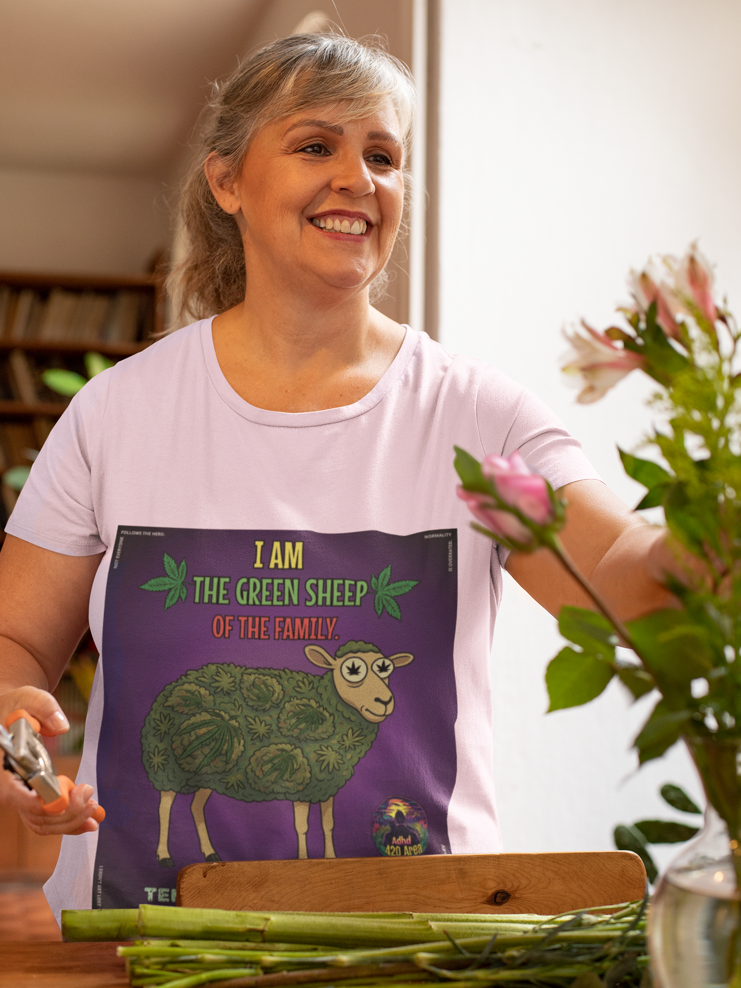 Eine lächelnde, ältere Frau mit grau-blonden Haaren arrangiert einen Blumenstrauß. Sie trägt ein hellviolettes T-Shirt von Temple of Green Elixir mit dem 'I am the Green Sheep of the Family'-Aufdruck. Die Szene ist hell und freundlich.﻿