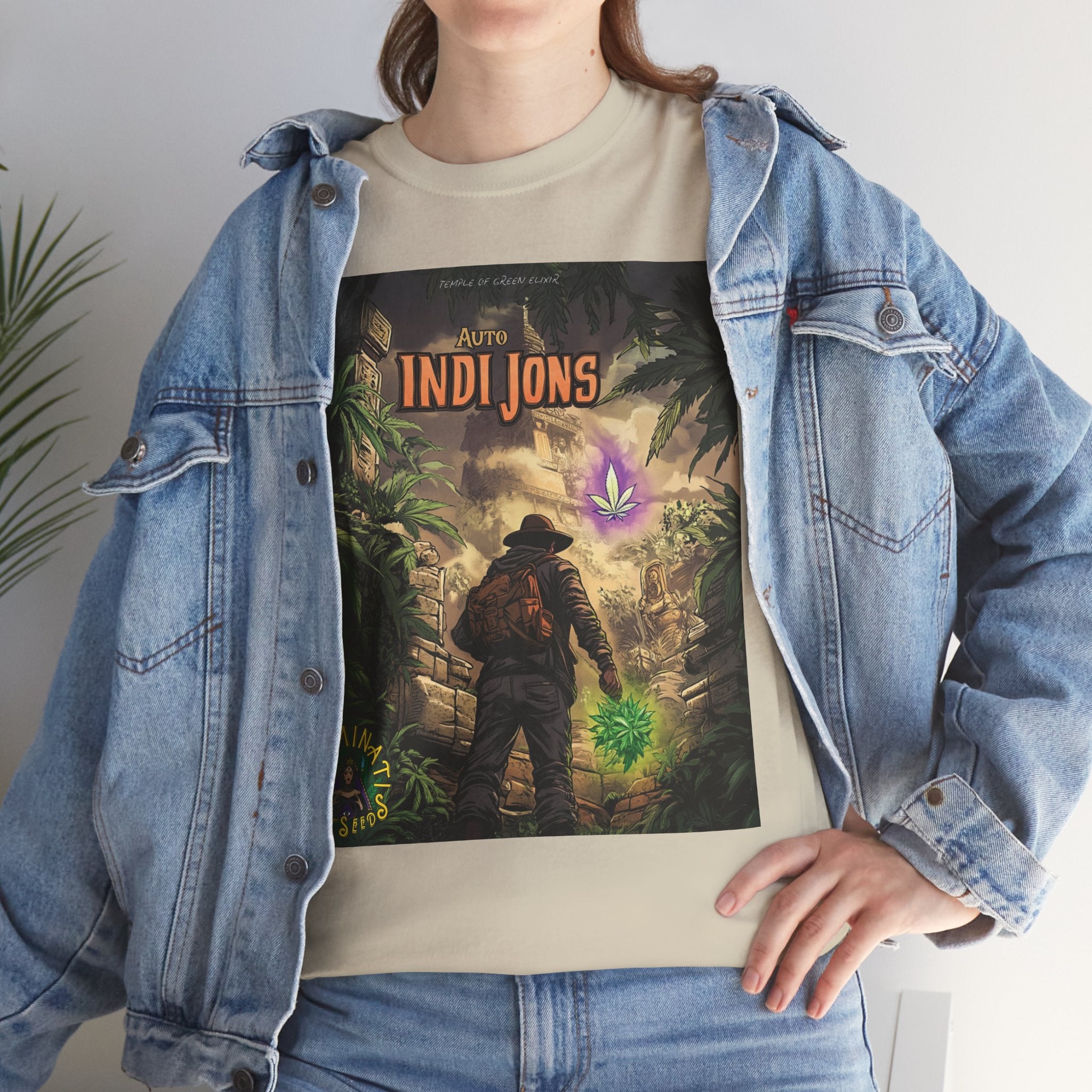 Frau trägt ein beiges T-Shirt mit dem „Auto Indi Jons“-Motiv von Illuminati Lost Seeds und Temple of Green Elixir. Das Design zeigt einen Abenteurer im Dschungel, der einem leuchtenden Cannabisblatt in einer antiken Tempelruine folgt. Über dem T-Shirt trägt sie eine offene Jeansjacke – eine Kombination aus urbanem Stil und mystischer Abenteuer-Ästhetik.