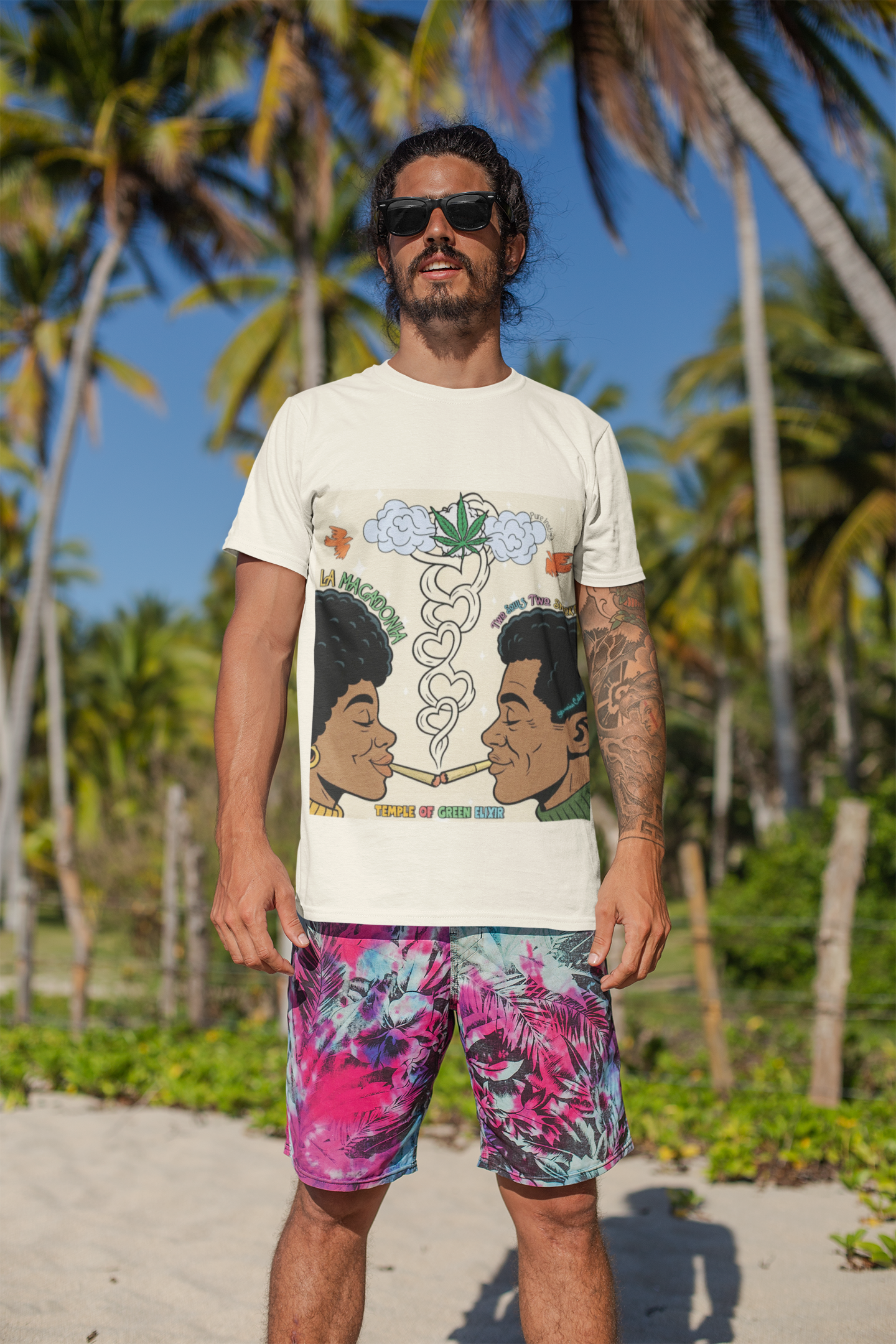 Mann steht am Strand vor Palmen, trägt ein weißes T-Shirt mit bunter Grafik von zwei Personen, die miteinander rauchen, Rauch in Herzform, Cannabisblatt und englischem Text „La Macdonia”, „The Souls Type Smoke”, „Temple of Green Elixir”, dazu bunte Shorts mit Blattmuster.