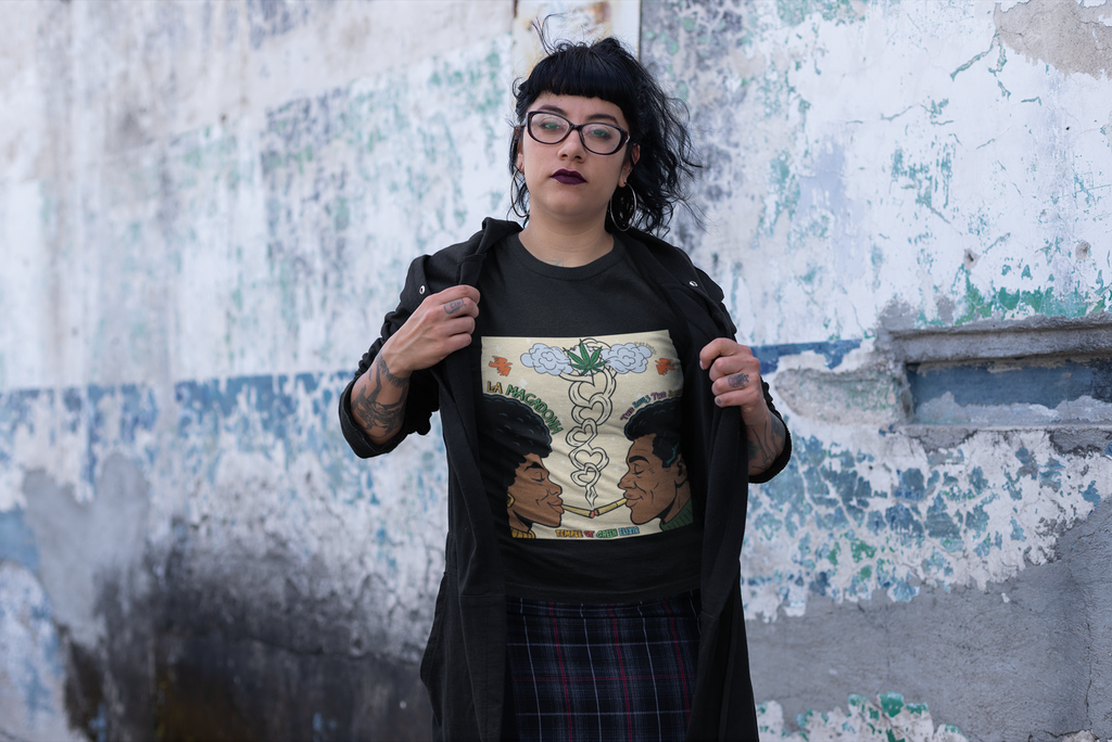 Person zeigt ein schwarzes T-Shirt mit bunter Grafik: Zwei Menschen rauchen gemeinsam, Rauch steigt in Herzform nach oben zu einer Cannabiswolke, umgeben von den Texten „La Macdonia“, „The Souls Type Smoke“ und „Temple of Green Elixir“. Im Hintergrund ist eine verwitterte Wand.