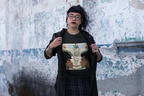 Person zeigt ein schwarzes T-Shirt mit bunter Grafik: Zwei Menschen rauchen gemeinsam, Rauch steigt in Herzform nach oben zu einer Cannabiswolke, umgeben von den Texten „La Macdonia“, „The Souls Type Smoke“ und „Temple of Green Elixir“. Im Hintergrund ist eine verwitterte Wand.