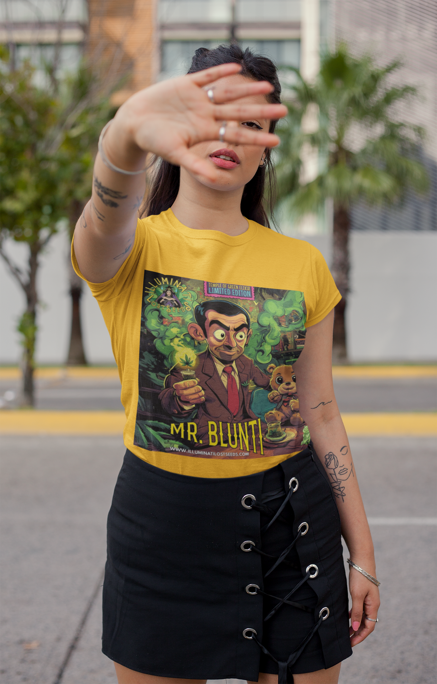 Junge Frau hebt ihre Hand vor das Gesicht, trägt gelbes  Mr. Blunti T-Shirt und schwarzen Rock auf der Straße. Auf dem T-shirt steht ein Mann im Anzug hält einen brennenden Joint und sitzt neben einem lächelnden Teddybär in rauchgeschwängerter, grüner Umgebung. Oben steht ‚Temple of Green Elixir Limited Edition‘, unten der Schriftzug ‚MR. BLUNTI‘ und das Illuminati Lost Seeds Logo.