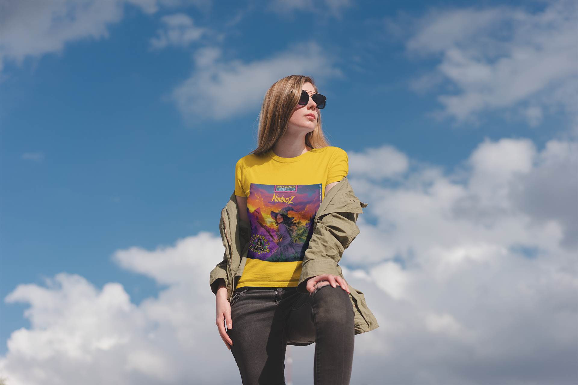 Junge braunharige Dame steht vor dem Himmel in Sonnenbrille und NimbusZ gelbes T-shirt mit Motiv: Hexe im violetten Zauberergewand mit Hut steht mit einem Stab in der Hand in einer Landschaft bei Sonnenuntergang, zwischen grünen Cannabis-Pflanzen. Oben der Schriftzug ‚NimbusZ‘ und ‚Temple of Green Elixir Limited Edition‘, unten das Logo ‚Illuminati Lost Seeds‘.