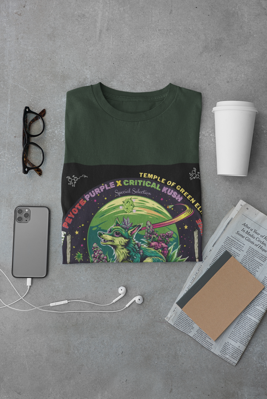Peyote Critical T-shirt grün zusammengefalten zwischen Brille, Handy, Zeitung, Becher. Auf dem T-shirt Ein smaragdgrün bepelzter Wolf patrouilliert zwischen lilafarbenen cannabis Blüten im Mondlicht und beobachtet jede Regung auf dem Feld. Sein Fell passt sich den Schattierungen der umliegenden Pflanzen an, als wäre er selbst ein verborgenes Geschöpf der Natur.