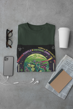 Peyote Critical T-shirt grün zusammengefalten zwischen Brille, Handy, Zeitung, Becher. Auf dem T-shirt Ein smaragdgrün bepelzter Wolf patrouilliert zwischen lilafarbenen cannabis Blüten im Mondlicht und beobachtet jede Regung auf dem Feld. Sein Fell passt sich den Schattierungen der umliegenden Pflanzen an, als wäre er selbst ein verborgenes Geschöpf der Natur.