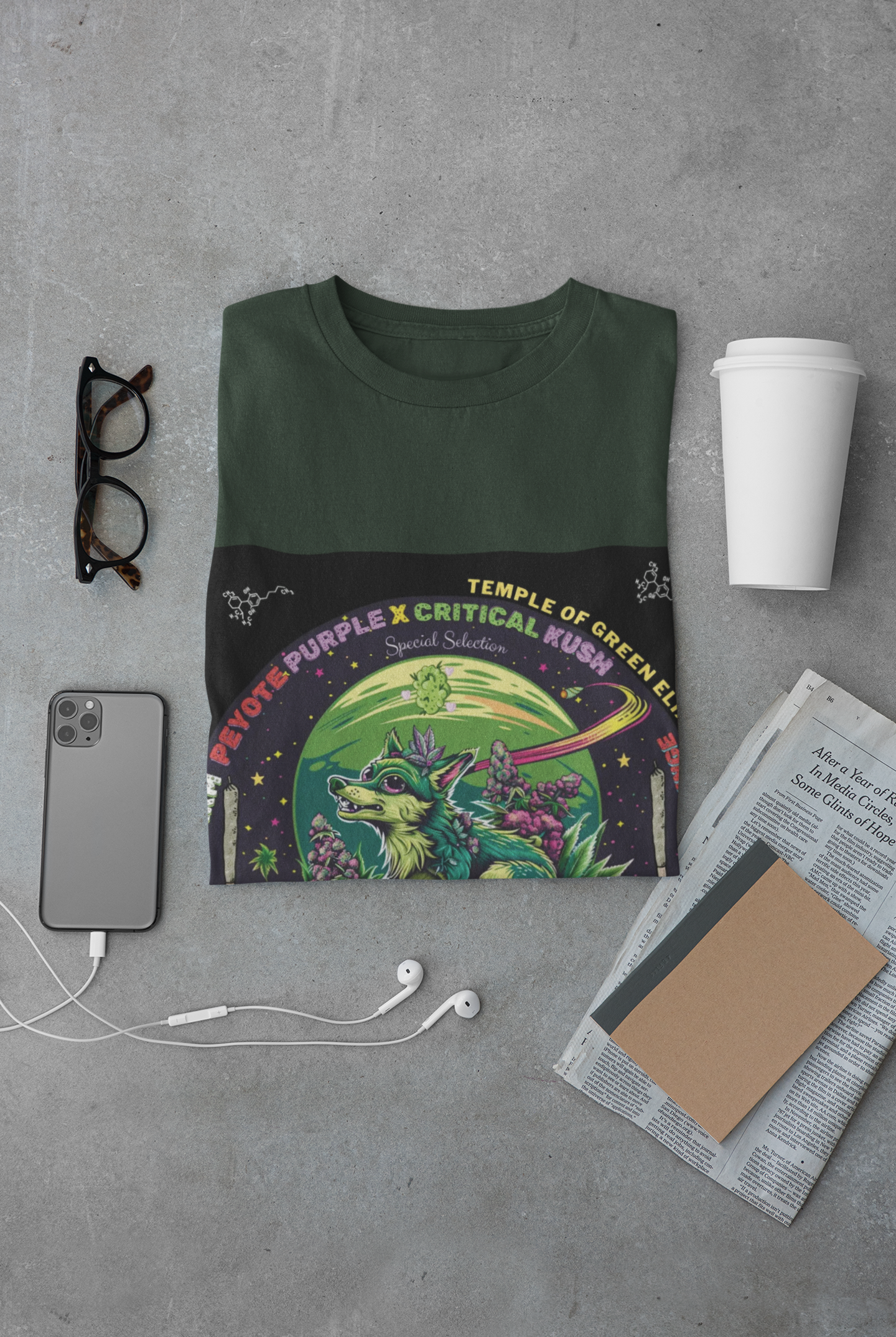 Peyote Critical T-shirt grün zusammengefalten zwischen Brille, Handy, Zeitung, Becher. Auf dem T-shirt Ein smaragdgrün bepelzter Wolf patrouilliert zwischen lilafarbenen cannabis Blüten im Mondlicht und beobachtet jede Regung auf dem Feld. Sein Fell passt sich den Schattierungen der umliegenden Pflanzen an, als wäre er selbst ein verborgenes Geschöpf der Natur.