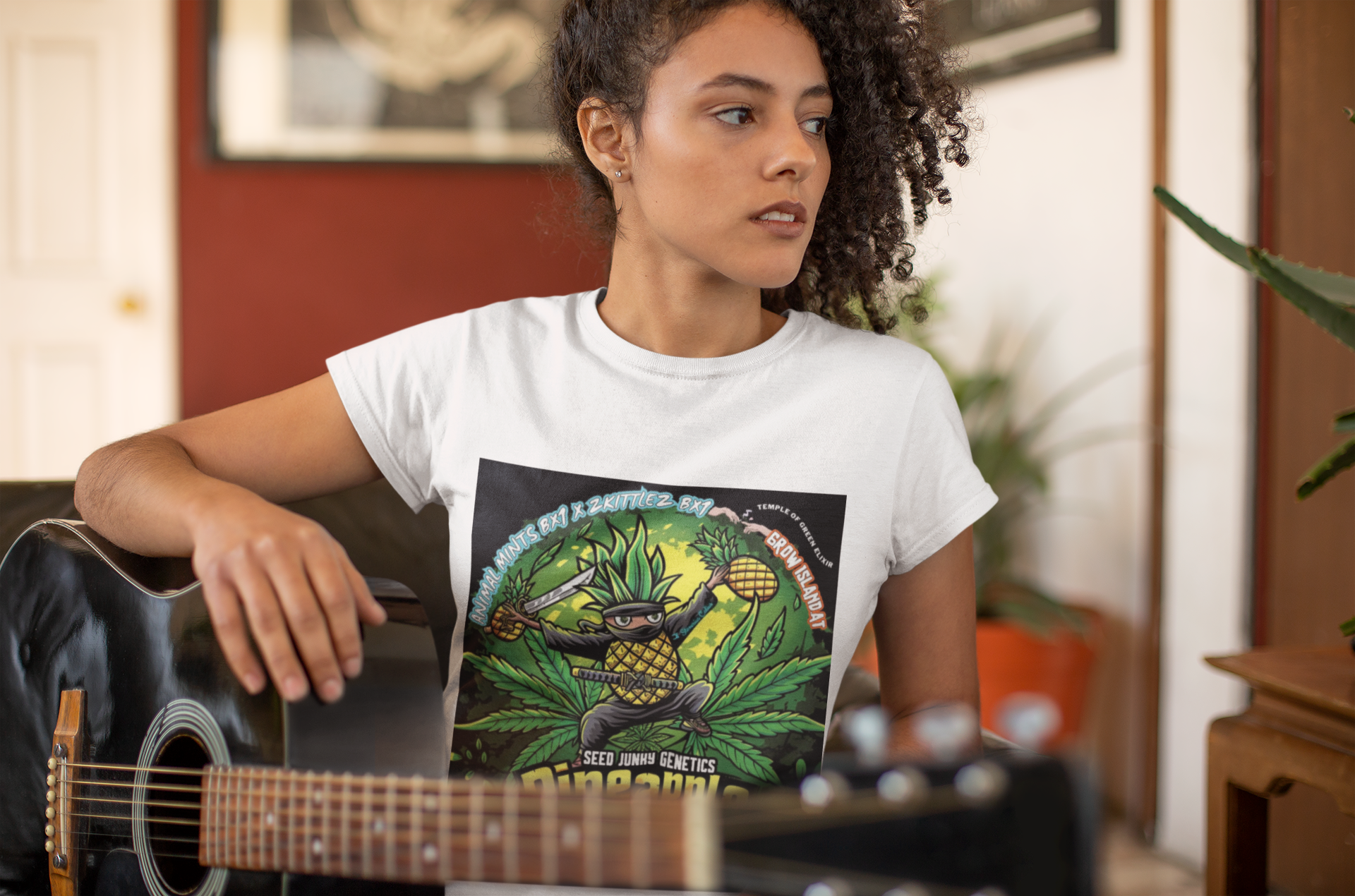 Dame mit lockigem Haar, das Gitarre spielt, trägt ein weißes Pineapple Fruz T-Shirt. Auf dem T-Shirt lässige Ananas-Ninja-Figur mit einem leuchtenden Katana und hoch erhobener frischen Ananas im Hintergrund sind üppige Cannabisblätter zu sehen.