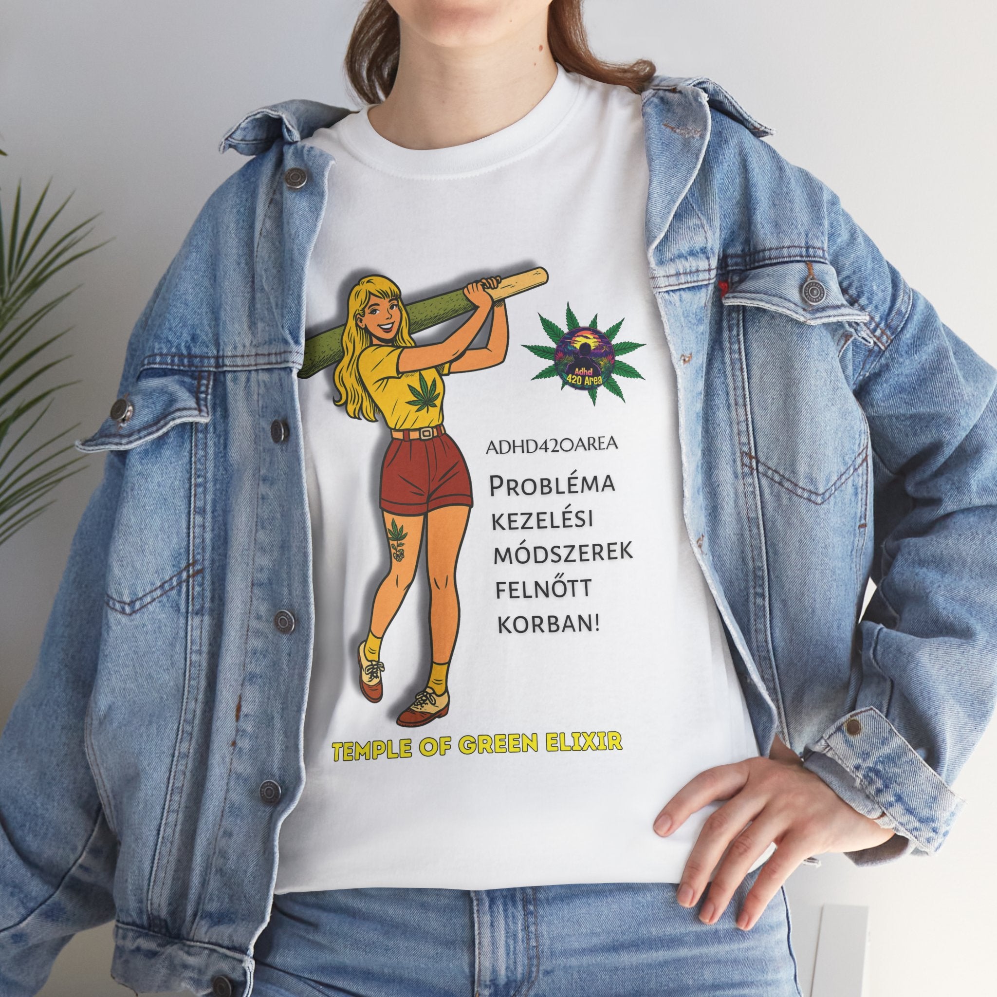 Junge Frau trägt ein weißes T-Shirt mit einer Grafik einer gezeichneten Figur, die eine große grüne Zigarette hält. Daneben sind Hanfblätter und der Text auf Deutsch: „ADHD42QAREA Problembehandlungsmethoden im Erwachsenenalter! Temple of Green Elixir“. Sie steht vor weißem Hintergrund und trägt eine blaue Jeansjacke.