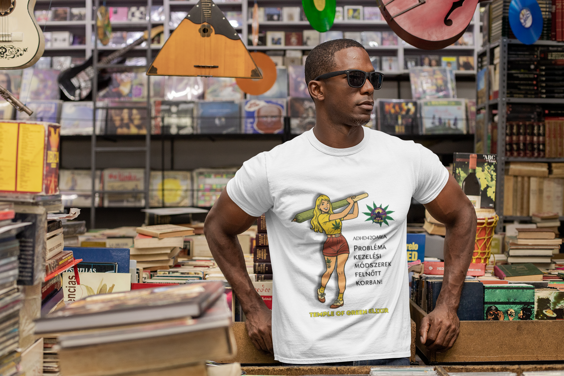 Mann steht in einem Plattenladen zwischen Bücherregalen und trägt ein weißes T-Shirt mit einer Zeichnung einer Frau mit einem großen grünen Joint. Auf dem Shirt ist ein Hanfblatt und der deutsche Text „ADHD42QAREA Problembehandlungsmethoden im Erwachsenenalter! Temple of Green Elixir“ zu sehen.