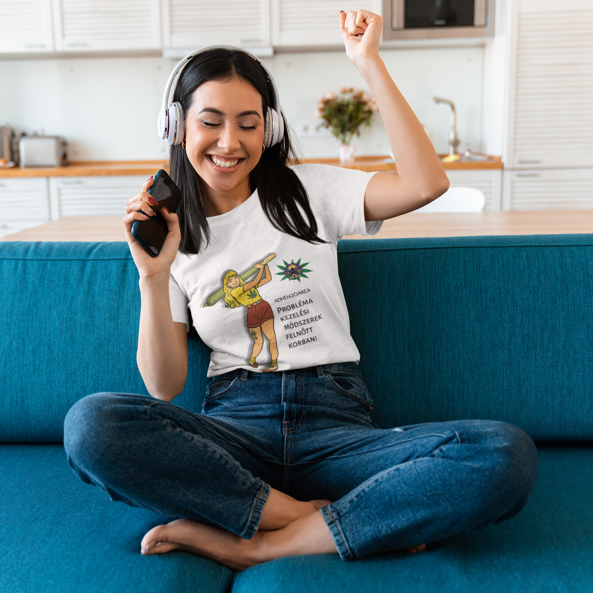 Frau sitzt entspannt auf einem blauen Sofa in einer modernen Küche, trägt ein weißes T-Shirt mit Zeichnung einer Frau, die einen großen grünen Joint hält. Das T-Shirt zeigt ein Hanfblatt und den deutschen Text „ADHD42QAREA Problembehandlungsmethoden im Erwachsenenalter! Temple of Green Elixir“. Die Frau hat Kopfhörer auf und hält ein Smartphone in der Hand.