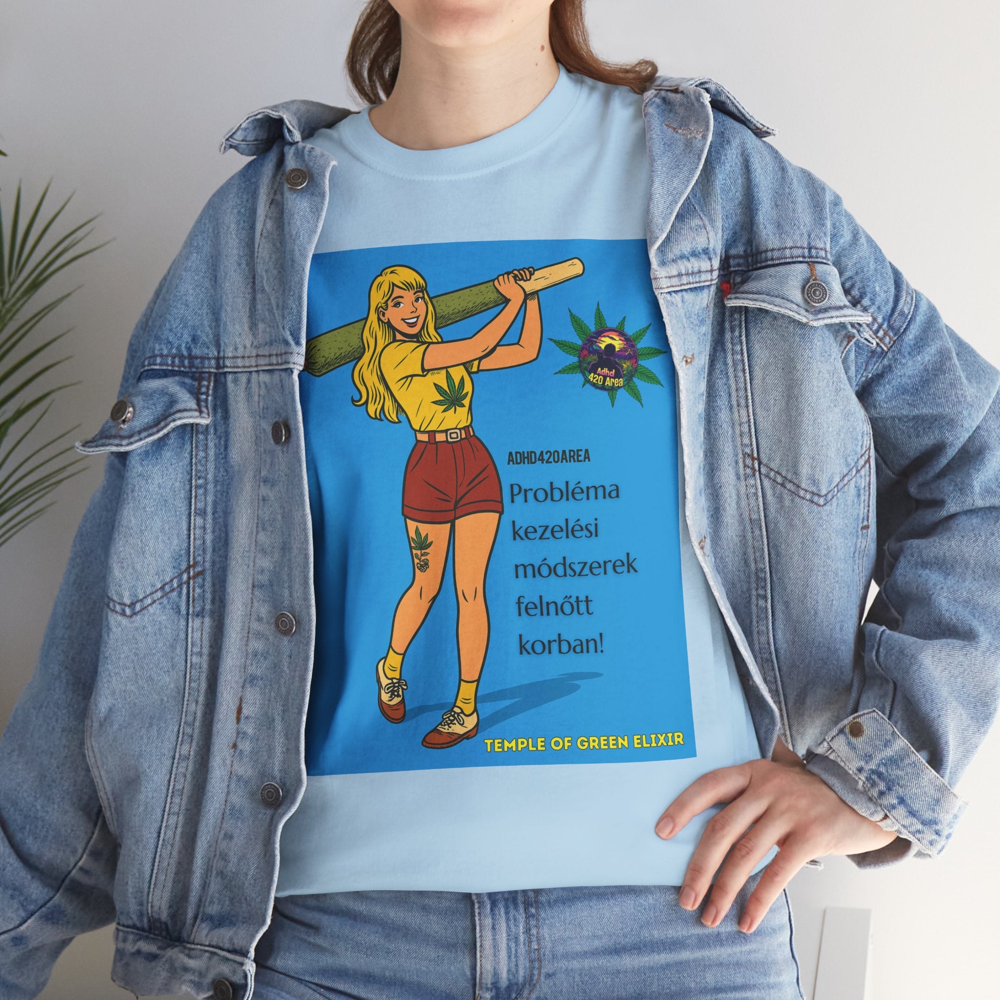 Junge Frau trägt ein blaues T-Shirt mit einer Grafik einer gezeichneten Figur, die eine große grüne Zigarette hält. Daneben sind Hanfblätter und der Text auf Deutsch: „ADHD42QAREA Problembehandlungsmethoden im Erwachsenenalter! Temple of Green Elixir“. Sie steht vor blauem Hintergrund und trägt eine blaue Jeansjacke.