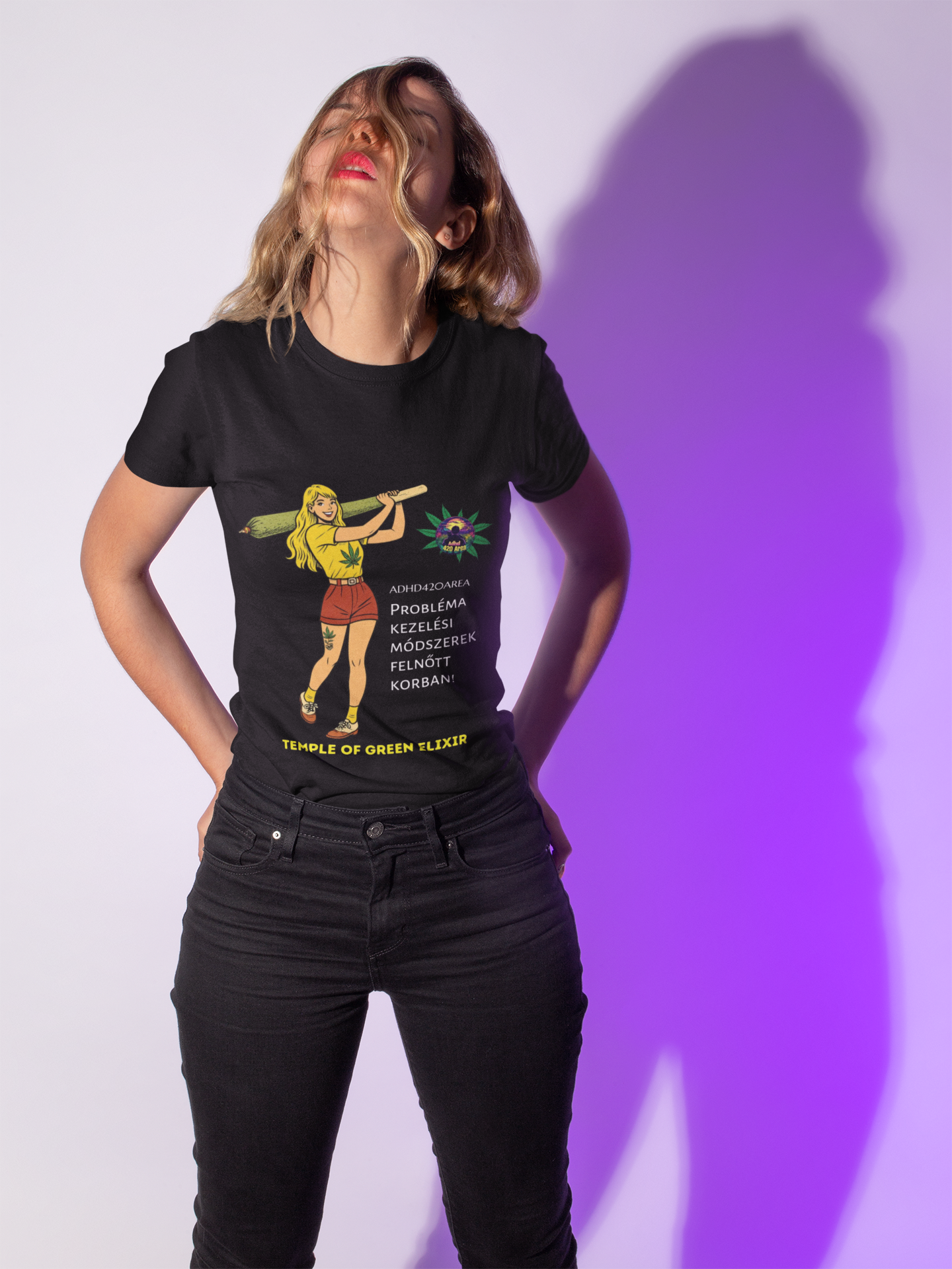 Frau trägt ein schwarzes T-Shirt mit einer bunten Grafik einer Frau, die einen großen grünen Joint hält, sowie Hanfblatt-Design und dem deutschen Text „ADHD42QAREA Problembehandlungsmethoden im Erwachsenenalter! Temple of Green Elixir“. Sie steht locker vor weißem Hintergrund mit lila Lichtschatten.