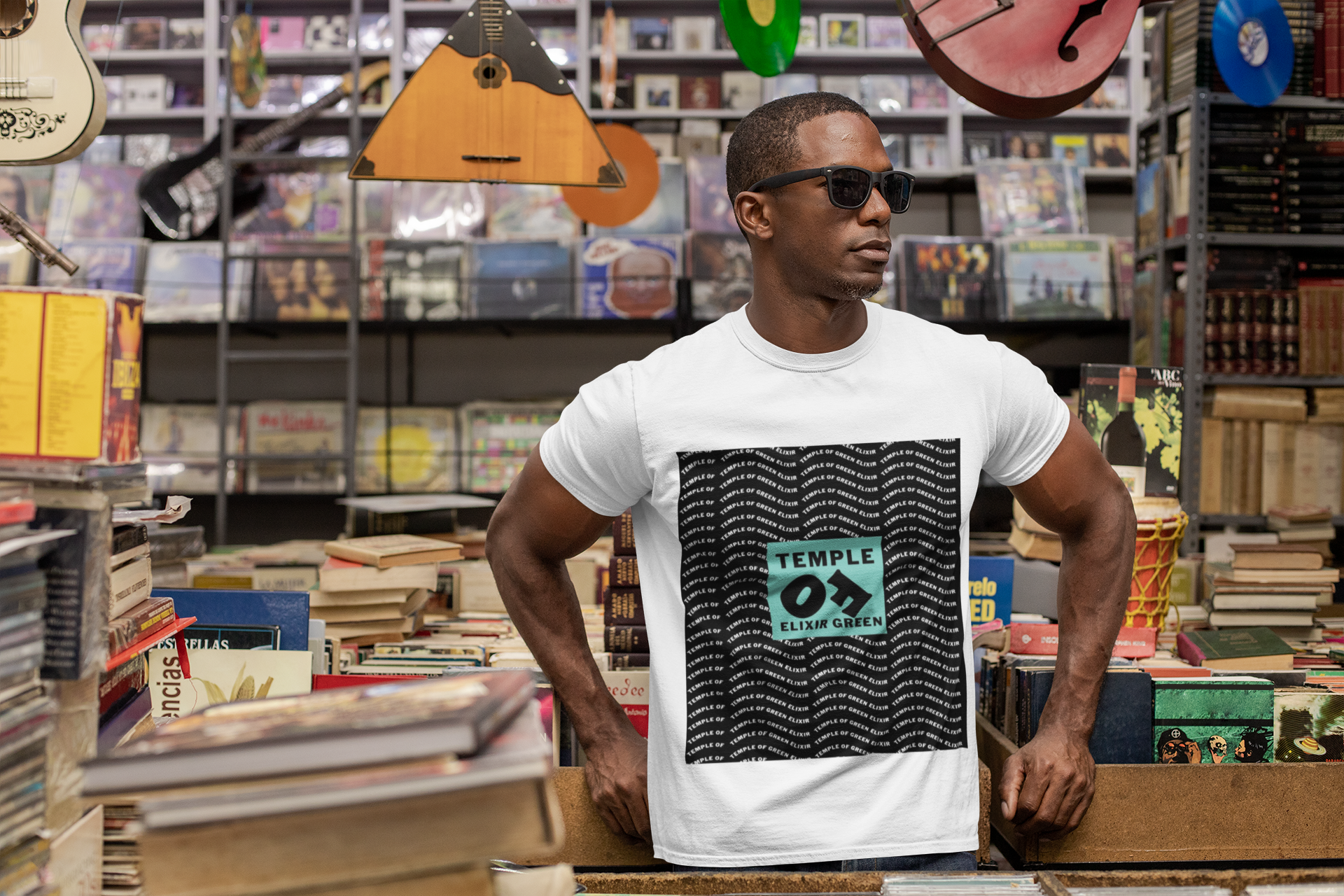 Ein Mann steht und lehnt sich in einem Plattenladen vor CDs und Büchern an. Er trägt eine schwarze Sonnenbrille und ein weißes Signature Pattern Tee T-Shirt. Schwarzer Hintergrund mit einer fließenden, wiederholenden Logostruktur, in der Mitte ein ikonischer türkisfarbener Block mit Temple of Green Elixir Schrift.