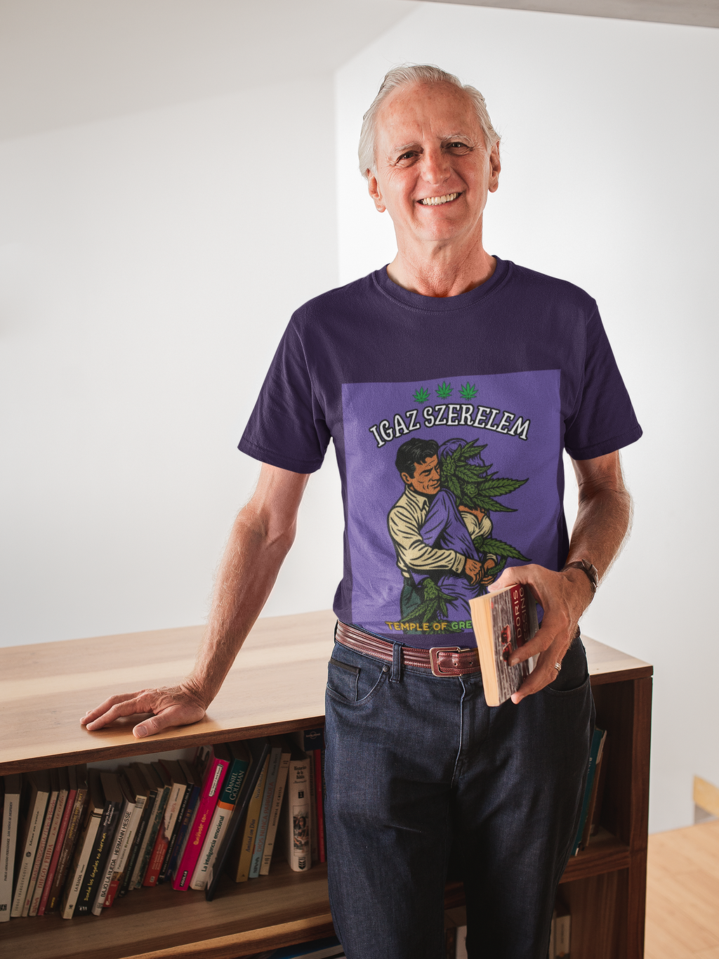 Ein eleganter, älterer Herr mit weißem Haar lehnt lässig an einem Bücherregal und hält ein Buch in der Hand. Er trägt das dunkelviolette T-Shirt mit dem ironischen 'Igaz Szerelem'-Aufdruck und beweist mit seinem Lächeln, dass Humor und ein guter Geschmack kein Alter kennen.﻿