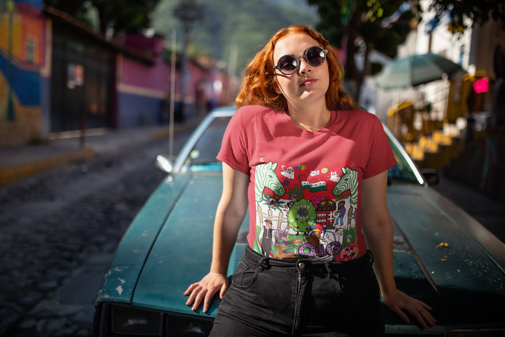 Eine junge Frau mit roten, lockigen Haaren und runder Sonnenbrille lehnt lässig an der Motorhaube eines blaugrünen Oldtimers. Sie trägt ein rotes Weltfrieden T-Shirt von Temple of Green Elixir mit einem bunten, satirischen Cartoon-Druck, der ungarische Motive thematisiert. Die Szene spielt auf einer Straße mit farbenfrohen Häusern im Hintergrund.