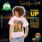 Lockige Dame lächelt, schaut nach links neben sich und richtet mit der rechten Hand ihr rosa Super Lemon Haze T-Shirt. Auf dem T-shirt die Grüne Lady – hebt langsam eine Zitronenscheibe an ihre Lippen, als würde sie den Sonnenschein selbst kosten. von ihr nach links schöne Cannabis blättern