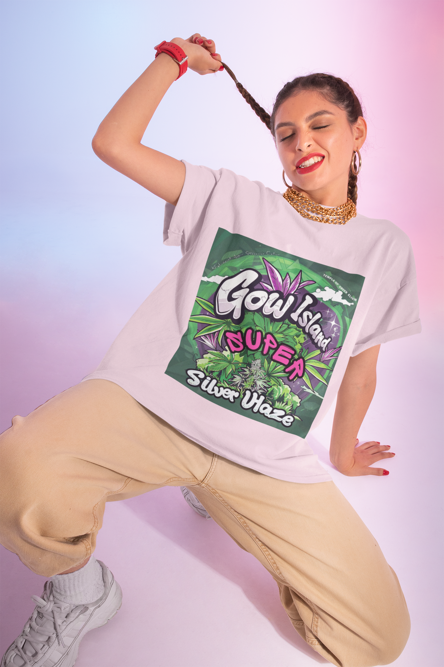 Eine Frau spielt mit ihren lockigen Haaren, zwinkert mit geschlossenen Augen. Sie trägt ein rosa  Super Silver Haze T-Shirt und beige Hose, vor einem rosa Hintergrund. Auf dem T-Shirt Leuchtende Grün- und Lilatöne, detailreiche Cannabis-Pflanzenmotive und markante Typografie.