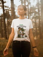 Eine Frau steht mit geschlossenen Augen im Wald und genießt den Moment der Ruhe. Sie trägt ein weißes T-Shirt mit der Aufschrift „IGAZ SZERELEM“ und einer Illustration, auf der eine Person liebevoll eine große Cannabispflanze umarmt. Darunter steht der Text „Temple of Green Elixir“. Die Sonne scheint sanft durch die Bäume, wodurch ein warmer, goldener Lichtschein entsteht. Das Bild vermittelt Frieden, Natürlichkeit und Achtsamkeit.