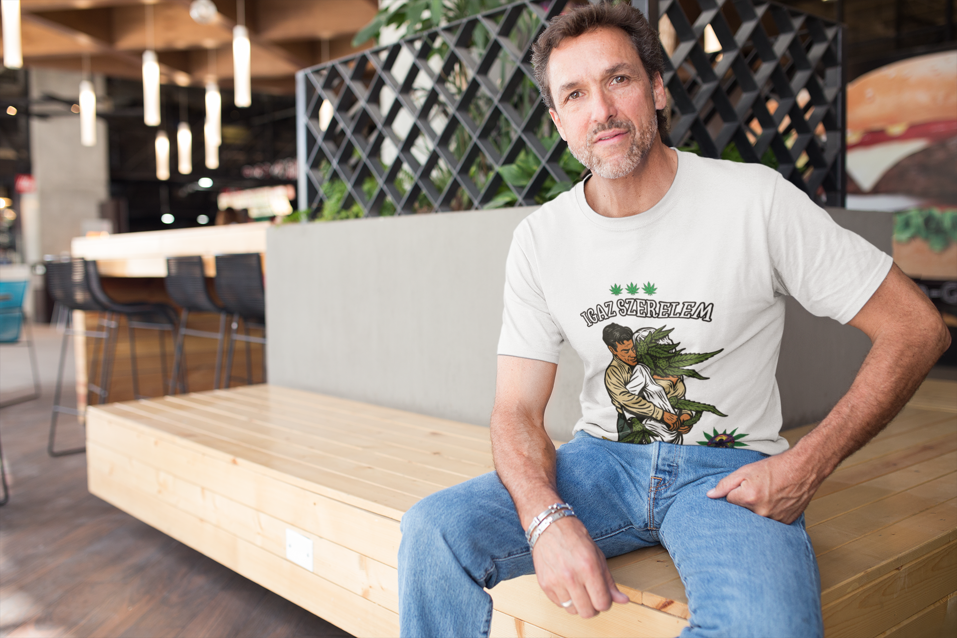 Ein Mann sitzt entspannt auf einer hellen Holzbühne in einem modernen Café oder Restaurant. Er trägt ein weißes T-Shirt mit der Aufschrift „IGAZ SZERELEM“ und einer Illustration, auf der eine Person eine große Cannabispflanze umarmt. Über der Grafik sind drei kleine Hanfblätter zu sehen. Der Mann schaut freundlich in die Kamera, im Hintergrund befinden sich Tische, Stühle und hängende Lampen. Die Szene wirkt ruhig, natürlich und stilvoll.