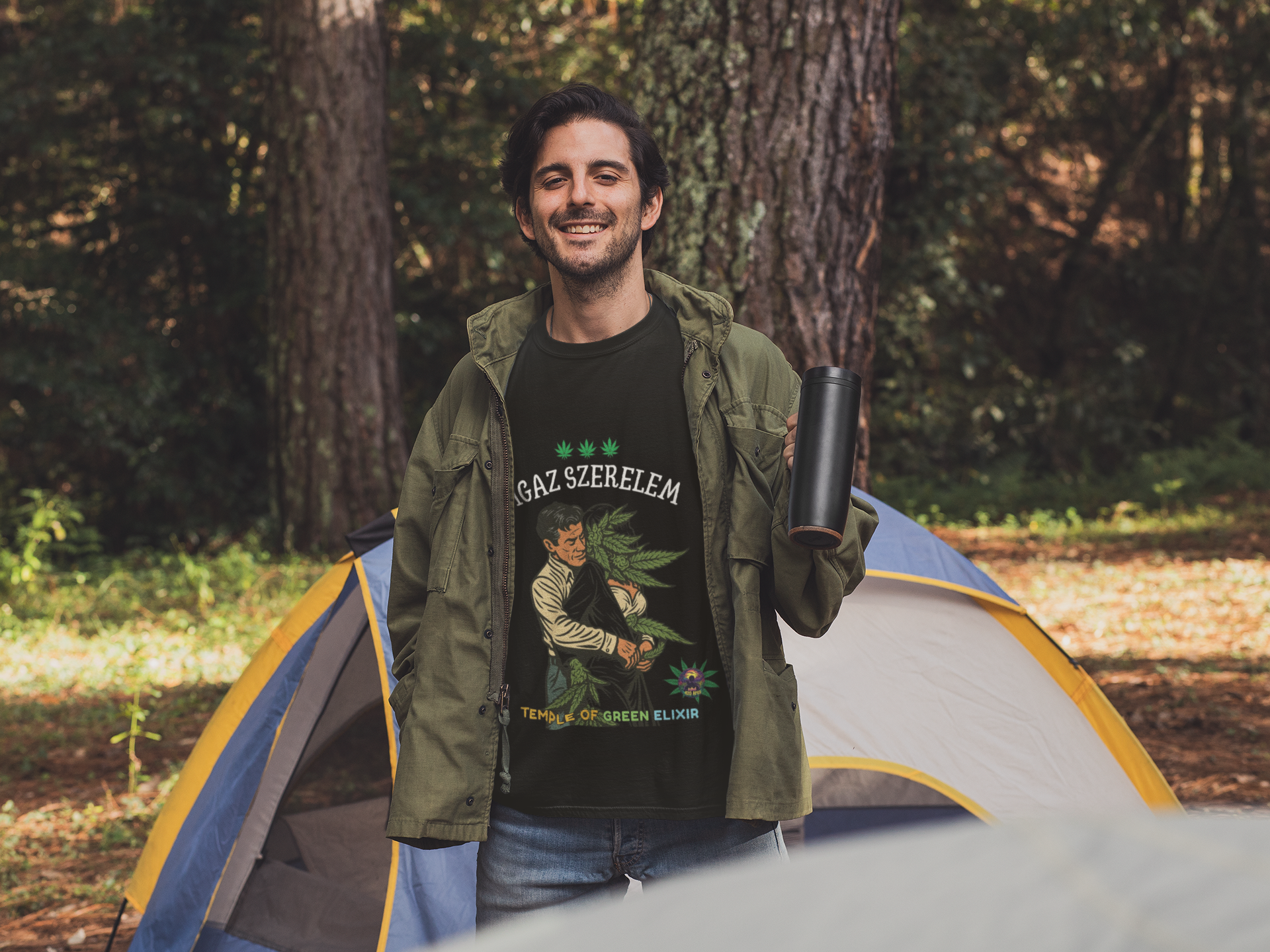 Ein Mann steht lächelnd vor einem Zelt im Wald und hält einen schwarzen Thermobecher in der Hand. Er trägt ein dunkles T-Shirt mit der Aufschrift „IGAZ SZERELEM – TEMPLE OF GREEN ELIXIR“ und einer Illustration, die eine Person zeigt, die eine große Cannabispflanze umarmt. Der Mann trägt eine grüne Outdoorjacke und Jeans, das Zelt im Hintergrund ist blau und gelb, und die Szene vermittelt ein natürliches, entspanntes Campinggefühl.