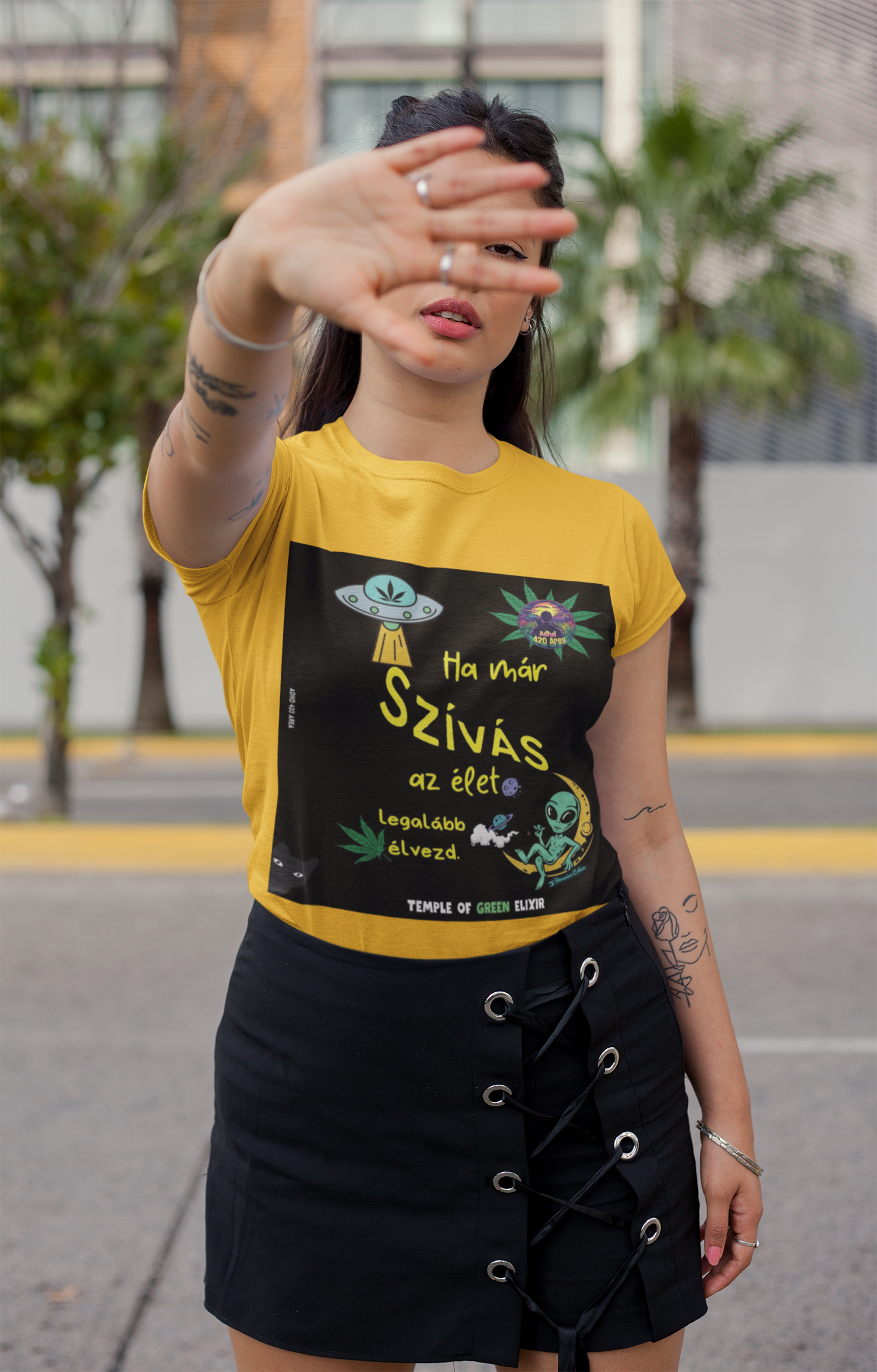 Frau trägt ein gelbes T-Shirt mit buntem Grafikdesign: UFO, Hanfblatt, rauchender Alien auf einer Mondsichel, ungarischem Text „Ha már szívás az élet, legalább élvezd. Temple of Green Elixir“, schwarzem Rock mit Schnürung, posiert draußen vor Hintergrund mit Palmen.