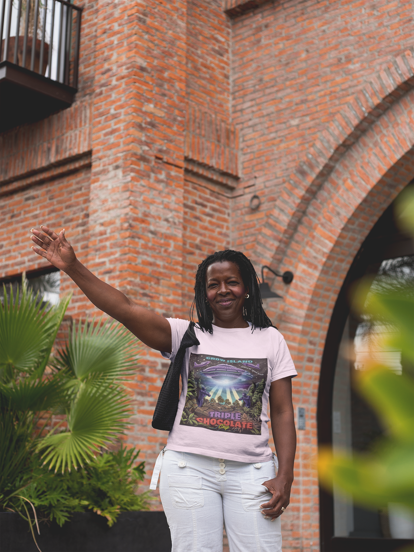 Eine Frau winkt, trägt ein rosa Triple Chocolate Chip T-Shirt, steht vor einer roten Backsteinmauer, links ist eine Palme. 
Im Zentrum der Illustration schwebt ein riesiges UFO, das mit einem grellen Lichtstrahl einen gigantischen Schokoriegel an Bord befördert – während futuristisch gerüstete Wächter das Gebiet sichern. Rundherum breiten sich üppige Cannabisfelder aus, im Hintergrund funkeln Sterne und zwei Raumschiffe vollenden die Szene.