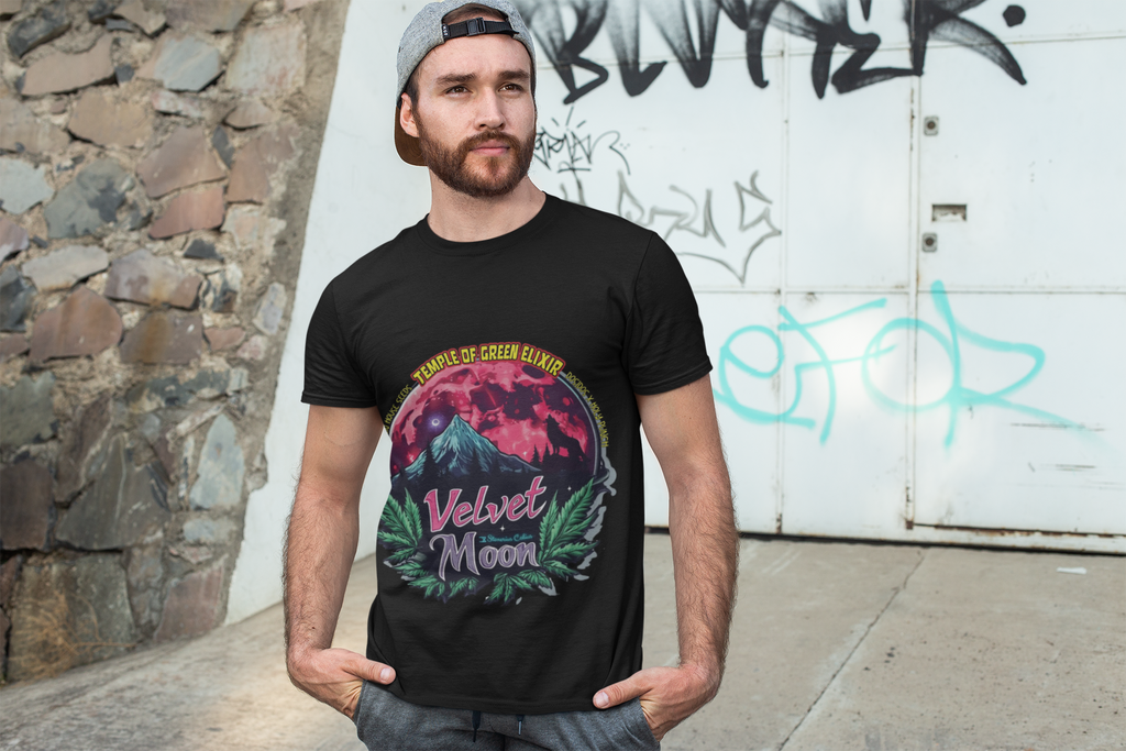 Ein Mann steht mit den Händen in den Taschen vor einer Garage, trägt eine Baseballkappe und ein schwarzes  Velvet Moon T-Shirt. Die Grafik erzählt von einer besonderen Nacht, in der das samtige Licht des blutroten Mondes die Berggipfel umhüllt und ein einsamer Wolf in den Schatten der dunklen Wälder hineinheult. Die Luft ist erfüllt vom Duft der frischen Bergluft, die die Freiheit der Wildnis und den Zauber des Unbekannten mit sich bringt. Cannabisblätter rahmen die Szene ein.