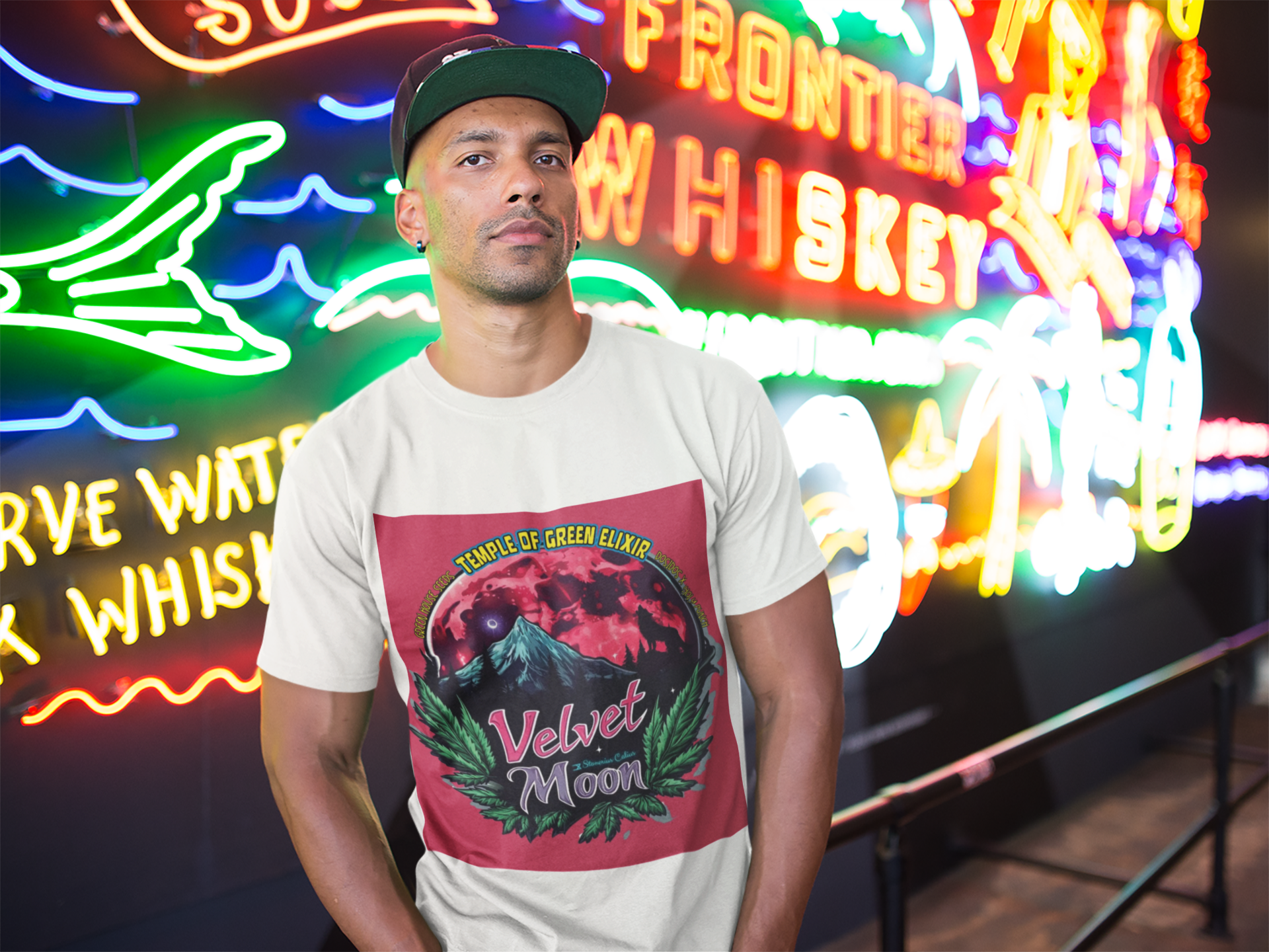 Ein junger Mann steht in einem weißen Velvet Moon T-Shirt mit einer Cap auf dem Kopf vor Neon-Schriften auf der Straße.  Die Grafik erzählt von einer besonderen Nacht, in der das samtige Licht des blutroten Mondes die Berggipfel umhüllt und ein einsamer Wolf in den Schatten der dunklen Wälder hineinheult. Die Luft ist erfüllt vom Duft der frischen Bergluft, die die Freiheit der Wildnis und den Zauber des Unbekannten mit sich bringt. Cannabisblätter rahmen die Szene ein.