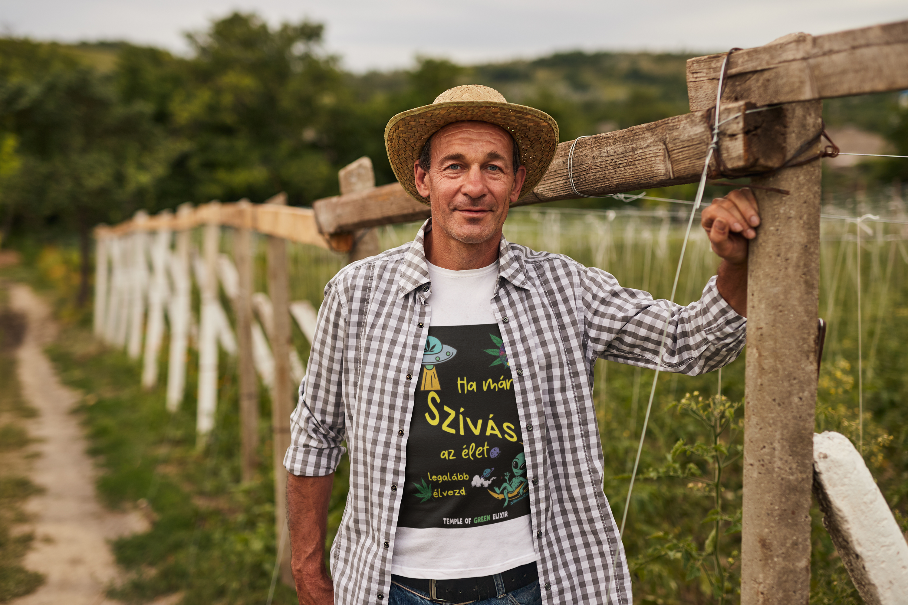 Mann mit Strohhut und kariertem Hemd steht an einem Holzpfosten auf einem Bauernhof, trägt ein weißes T-Shirt mit buntem Grafikdesign: UFO, Hanfblatt, rauchender Alien, ungarischer Text „Ha már szívás az élet, legalább élvezd. Temple of Green Elixir“, im Hintergrund ländliche Landschaft